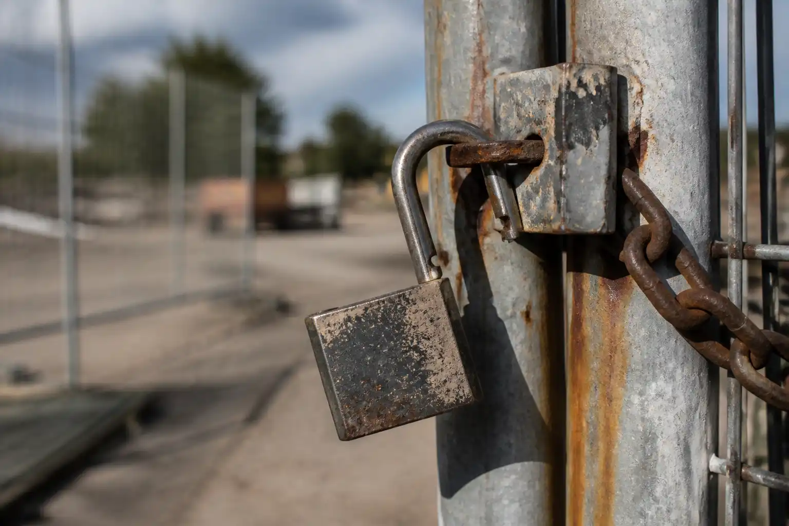 A heavy steel padlock hanging open on the hasp of a site gate, its shackle swung clear